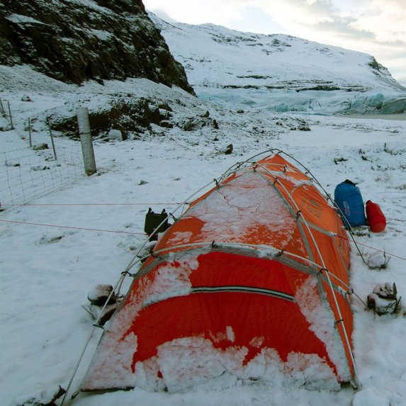 Et oransje ekspedisjonstelt, delvis dekket av sn, er satt opp i et vinterlig, steinete landskap med en isbre i bakgrunnen. Utenfor teltet ligger en bl og en rd BasicNature Dry Bag.