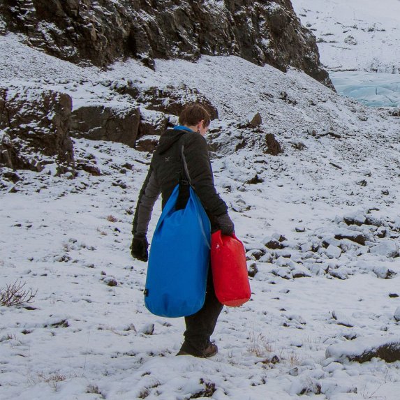 En person i mrke ytterklr gr i et sndekt, steinete landskap og brer to BasicNature Dry Bags: en stor, bl vanntett pakkpose over skulderen og en mindre, rd pakkpose i hnden, noe som demonstrerer deres bruk under tffe utendrsforhold.