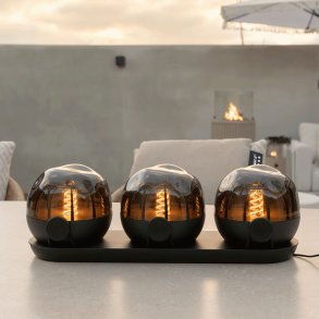 Kolme ladattavaa Cosisfera Black LED-tunnelmavalaisinta, asetettuna mustalle latausalustalle ulkopydll hmrss, takana nkyy ulkokeitti/terassiymprist kaasutakalla.