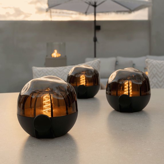 Kolme ladattavaa Cosisfera Black LED-tunnelmavalaisinta asetettuna suoraan valkoiselle ulkopydlle hmrss. Taustalla nkyy vaaleita tyynyj ja aurinkovarjo, ja mys kaasutakka on nkyviss.