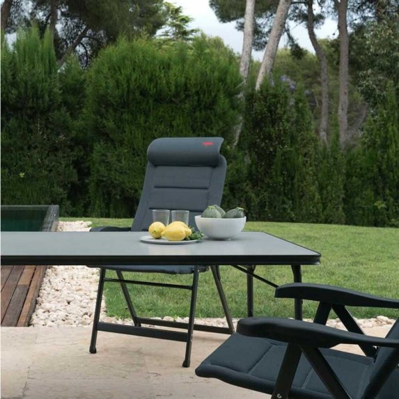 Luksuri�st og stilrent campingbord med gr� overflade og sort stel opstillet p� en terrasse i haven. Bordet er d�kket med citrusfrugter og sk�le, og i baggrunden ses en komfortabel campingstol, gr�spl�ne og pool. Velegnet som udend�rs spisebord.