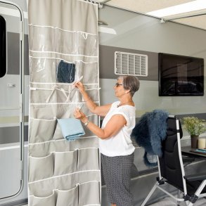 En kvinna i vit topp och mnstrad kjol hnger bestick och en bl trasa i en stor, hg Isabella Organizer Tall med ljusgr tygfickor och transparenta foliefickor. Organizeraren r monterad p sidan av en husvagn i ett frtlt.