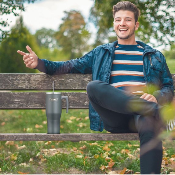 En ung mann sitter p en benk i en park, ifrt en bl jakke og en stripete genser. Han smiler og peker med hnden, mens han har en stor termoskop fra Thermos ved siden av seg p benken.